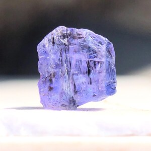 May include: A faceted, purple-blue gemstone with a rough, natural finish. The gemstone is translucent and has a slight sheen.