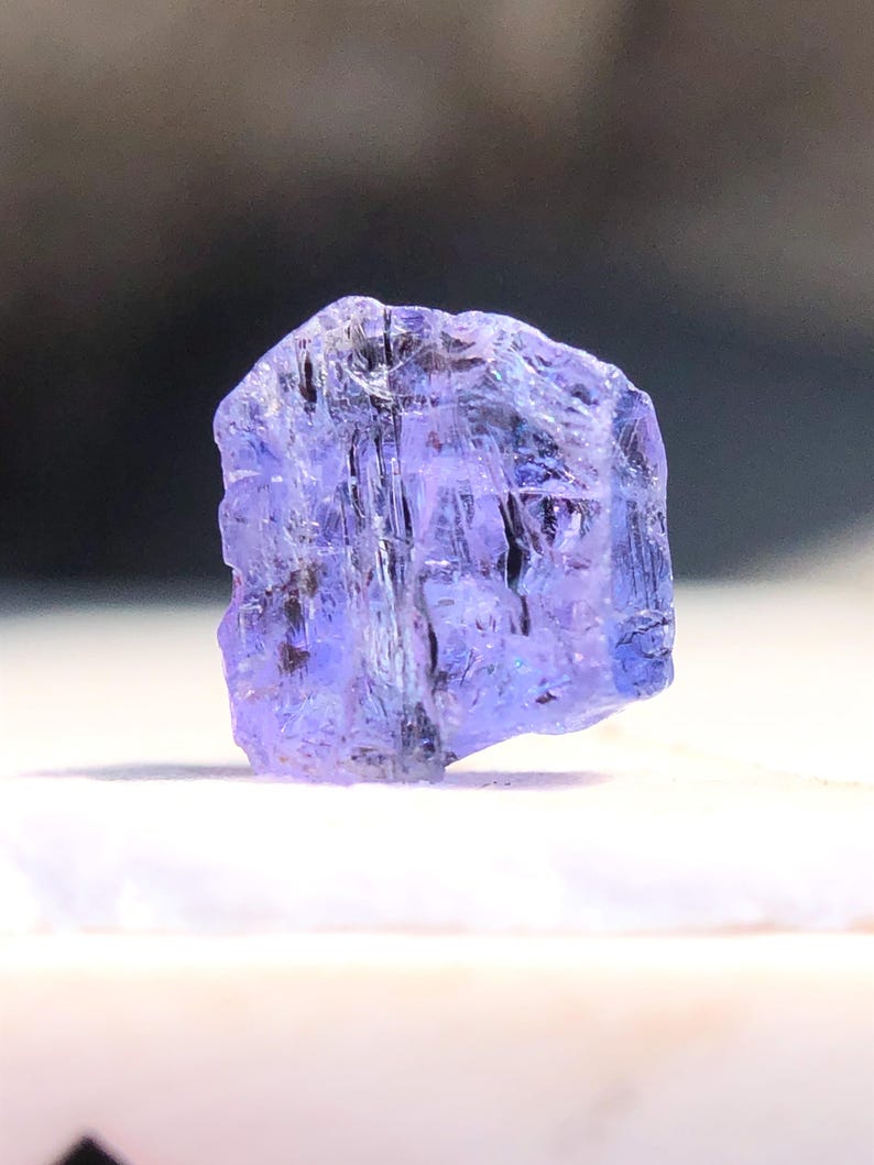 May include: A faceted, purple-blue gemstone with a rough, natural finish. The gemstone is translucent and has a slight sheen.