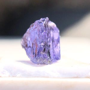 May include: A purple gemstone with a faceted, geometric shape. The crystal is translucent and has a slight sheen.
