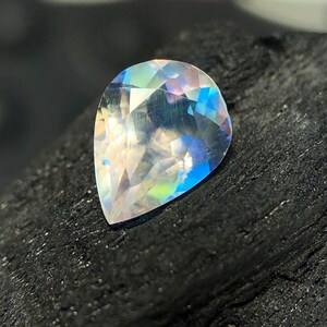 May include: A pear-shaped iridescent gemstone with a rainbow of colors, including blue, green, and pink. The gemstone is set against a dark background.