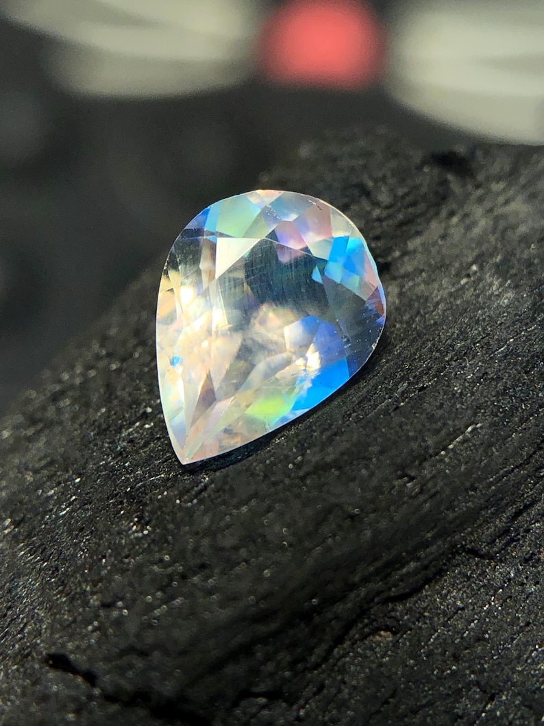 May include: A pear-shaped iridescent gemstone with a rainbow of colors, including blue, green, and pink. The gemstone is set against a dark background.