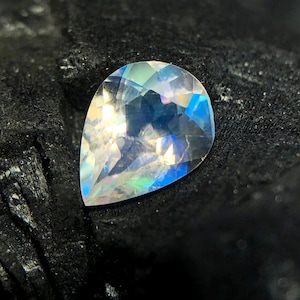 May include: A pear-shaped iridescent gemstone with a rainbow of colors, including blue, green, and pink. The gemstone is set against a black background.
