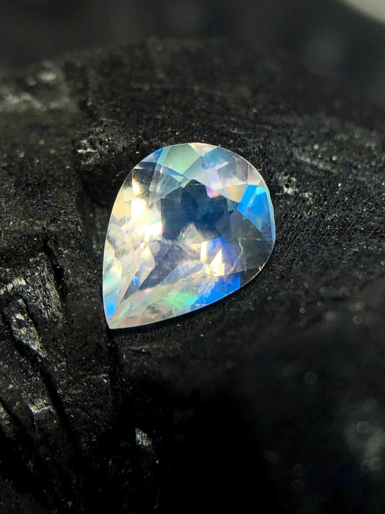 May include: A pear-shaped iridescent gemstone with a rainbow of colors, including blue, green, and pink. The gemstone is set against a black background.
