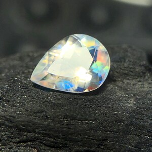 May include: A pear-shaped iridescent gemstone with a clear, faceted body. The gemstone is set against a dark, textured background.