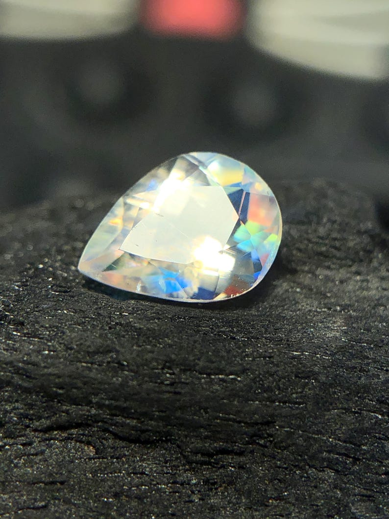 May include: A pear-shaped iridescent gemstone with a clear, faceted body. The gemstone is set against a dark, textured background.