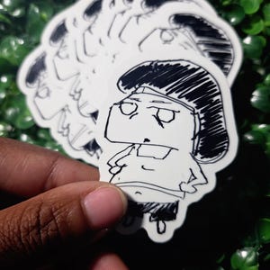 May include: A die-cut vinyl sticker featuring a black and white hand-drawn illustration of a cartoon character with a large head, dark hair, and a simple outfit. The character has a stern expression with hands on hips, rendered in a sketchy ink style.