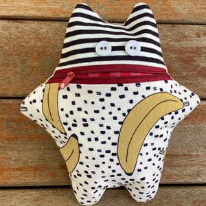 Könnte beinhalten: Ein Plüschtier in Form eines Monsters mit einer Reißverschlusstasche. Das Spielzeug ist aus weißem Stoff mit schwarzen Punkten und gelben Bananenmustern. Die Oberseite des Spielzeugs ist aus schwarz-weiß gestreiftem Stoff mit zwei weißen Knöpfen für die Augen.