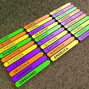 May include: A set of colorful wooden plant markers with the names of different herbs written on them. The markers are arranged in two rows, with the names of the herbs listed in alphabetical order. The markers are on a green grassy surface.