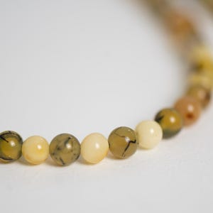 Op de afbeelding: Een close-up van een kralenketting met een verscheidenheid aan aardetinten. De kralen zijn voornamelijk geel, crème en bruin, sommige met donkere adering. De ketting is geplaatst tegen een witte achtergrond.