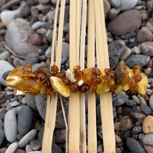Könnte beinhalten: Eine goldene Haarklammer, verziert mit Bernstein-Steinen in verschiedenen Formen und Größen. Die Steine sind eine Mischung aus hellem und dunklem Bernstein, einige haben ein trübes oder milchiges Aussehen. Die Klammer ist vor einem Hintergrund aus Natursteinen platziert.
