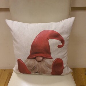 May include: A white cushion featuring a red and white gnome design. The gnome is wearing a red hat and has a white beard.