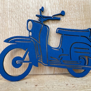 Könnte beinhalten: Blauer Holzausschnitt eines Vintage-Rollers. Der Roller hat eine abgerundete Front, einen Sitz und einen Lenker. Der Ausschnitt ist in einem einfarbigen Blau gehalten und vor einem hellen Holzgrund platziert.