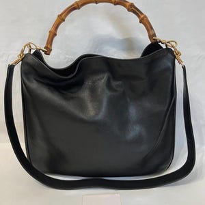 May include: A black leather hobo bag featuring a curved bamboo top handle and a detachable black leather shoulder strap with gold-tone metal hardware. A small white card with the text GUCCI is positioned in front of the bag.