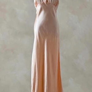 Könnte beinhalten: Ein pfirsichfarbenes, bodenlanges Slip-Kleid. Das Kleid hat einen quadratischen Ausschnitt mit gestickten Details und kleinen Knöpfen. Der Stoff hat einen seidigen Glanz und das Kleid hat einen leichten Schlag am Saum.