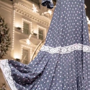 Op de afbeelding: Een lange, blauwe jurk met een wit bloemenpatroon en witte kanten afwerking. De jurk heeft een hoge halslijn, korte mouwen en een gelaagde rok. De achtergrond is een wazige afbeelding van een gebouw met kerstverlichting.