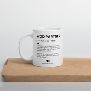Könnte beinhalten: Eine weiße Keramiktasse mit einem schwarzen Design, das einen "WOD Partner" als "Begleiter, der dir hilft, deine Fitnessziele zu erreichen" definiert. Die Tasse enthält auch den Hashtag #PARTNERWODFORLIFE.