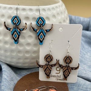 May include: Three pairs of wooden earrings with a geometric pattern. The earrings are shaped like a bull's head and are painted in blue, orange, and brown. The earrings are on a white card with the text "Gemini Creative".