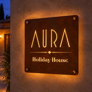Può includere: Un cartello rettangolare illuminato con la parola "AURA" in un carattere stilizzato, sopra "Holiday House". Il cartello è montato su un muro strutturato, con un'illuminazione calda sui bordi. Il cartello è marrone con lettere dorate.