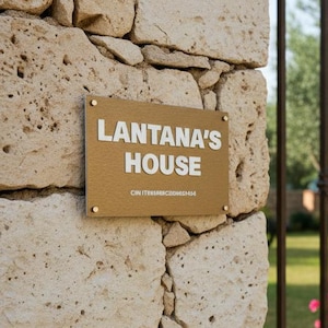 May include: A weathered stone wall with a gold sign reading "Lantana's House". A wrought iron gate is in the background, with a garden and pink flowers visible. The sign is fixed to the wall with small gold screws.