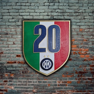 Puede incluir: Un letrero de madera con forma de escudo con los colores de la bandera italiana, verde, blanco y rojo. El número 20 está impreso en azul en la sección blanca. Un logotipo azul y blanco con las letras "NI" está en el centro del escudo.