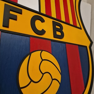Puede incluir: Un escudo de madera con el logotipo del FC Barcelona. El logotipo presenta las iniciales del equipo "FCB" en letras negras sobre fondo amarillo, un patrón de rayas rojas y amarillas, y un balón de fútbol negro y amarillo.