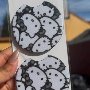 May include: Two car coasters with a white background and a repeating pattern of a cartoon cat face in gray and black. The coasters are packaged in a clear plastic sleeve. The text "Car Coasters Handmade with Love" is printed on the packaging.