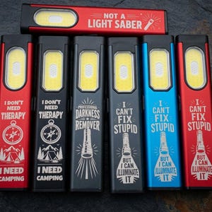 May include: Six rectangular LED flashlights in red, black, and blue with various engraved phrases including NOT A LIGHT SABER, I DON'T NEED THERAPY I NEED CAMPING, PROFESSIONAL DARKNESS REMOVER, and I CAN'T FIX STUPID BUT I CAN ILLUMINATE IT.
