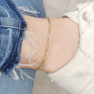 以下が含まれることがあります： 繊細なリンクデザインのゴールドチェーンのアンクレット。アンクレットは足首に着用されており、足首は白いスニーカーと青いジーンズの上に置かれています。