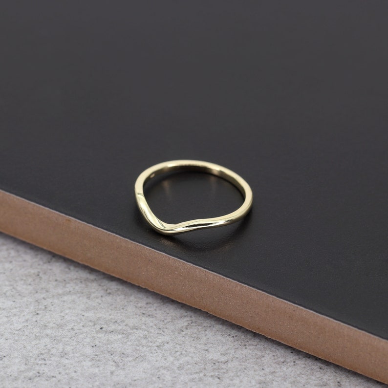 Op de afbeelding: Een eenvoudige gouden ring met een gebogen, v-vormig ontwerp. De ring ligt op een zwart oppervlak.