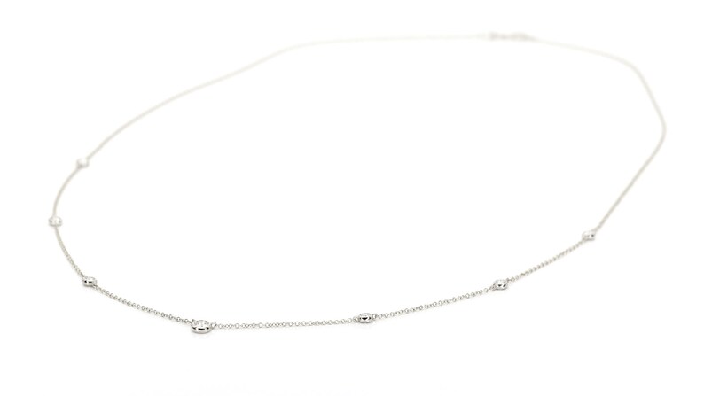 May include: A delicate silver necklace with several oval-shaped embellishments evenly spaced along the chain. The necklace is displayed against a white background, highlighting its minimalist design and elegant simplicity. The chain is thin and the embellishments are small.