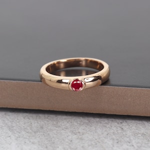 May include: A gold ring with a single, round, red gemstone set in the center. The ring has a smooth, polished band and a classic design. The gemstone is a vibrant red color, and the ring is displayed on a dark surface.