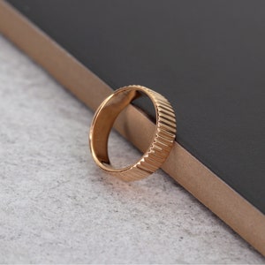 Puede incluir: Un anillo dorado con un dise&ntilde;o texturizado y estriado. El anillo est&aacute; sobre una superficie gris oscuro.