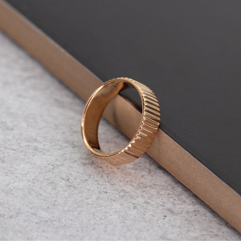 Puede incluir: Un anillo dorado con un dise&ntilde;o texturizado y estriado. El anillo est&aacute; sobre una superficie gris oscuro.