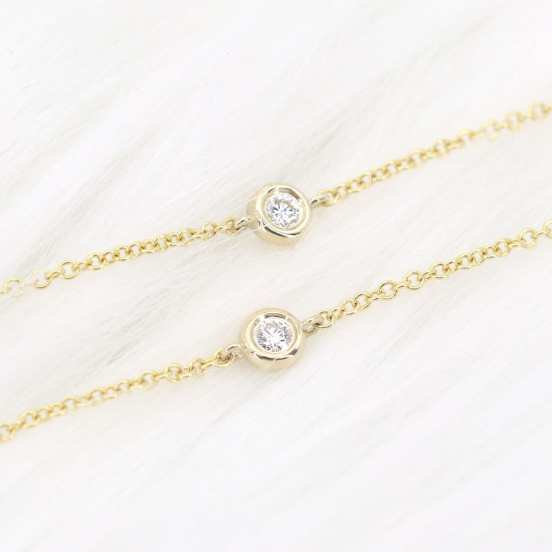 May include: Two delicate gold necklaces with bezel-set, round, clear gemstones. The necklaces are on a white, fluffy surface. The chains have a classic link design, and the gemstones are centered on the chains.