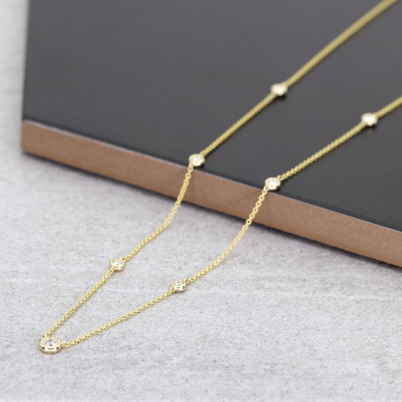 May include: A delicate gold necklace featuring multiple small, round, clear gemstones evenly spaced along the chain. The necklace is displayed on a dark gray surface, showcasing its elegant design and sparkle.