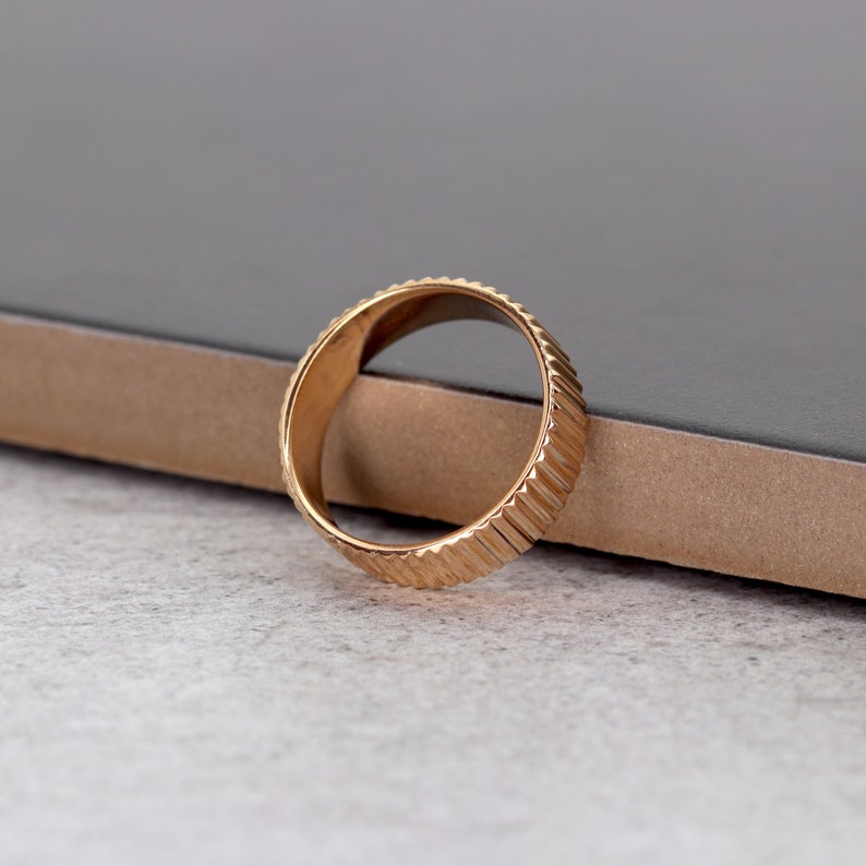 Puede incluir: Un anillo dorado con un dise&ntilde;o texturizado y estriado. El anillo est&aacute; sobre una superficie marr&oacute;n.