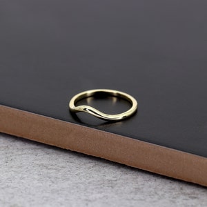 Op de afbeelding: Een eenvoudige gouden ring met een gebogen, golvend ontwerp. De ring wordt getoond op een zwart oppervlak.