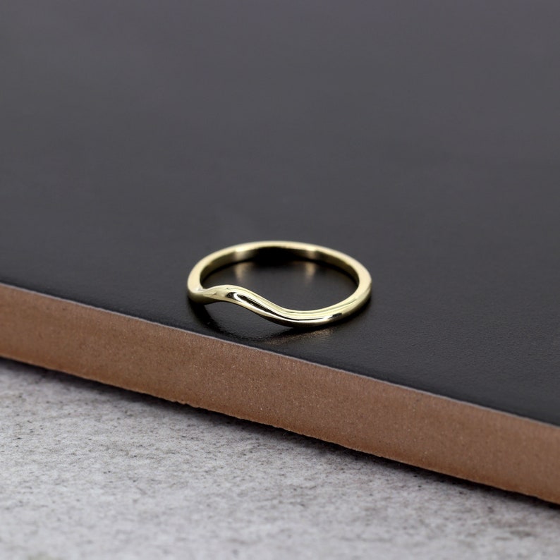 Op de afbeelding: Een eenvoudige gouden ring met een gebogen, golvend ontwerp. De ring wordt getoond op een zwart oppervlak.