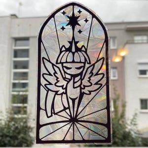 Puede incluir: Una pegatina de ventana de vidriera negra e iridiscente con un personaje de dibujos animados con alas, una corona y una estrella. El diseño está enmarcado en un marco negro arqueado, que se asemeja a una vidriera.