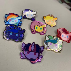 Könnte beinhalten: Eine Sammlung farbenfroher Emaille-Anstecknadeln mit Cartoon-Pony-Charakteren. Jede Anstecknadel hat ein einzigartiges Design mit leuchtenden Farben, darunter Blau, Lila, Rosa, Gelb und Orange. Die Anstecknadeln haben eine glänzende, metallische Oberfläche.