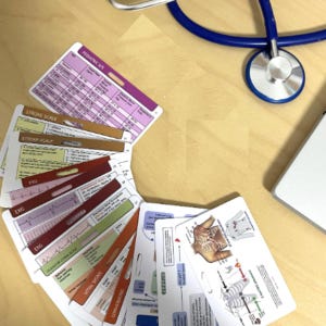 May include: A set of medical reference badge cards featuring color-coded charts for pediatric vital signs, stroke scales, EKG rhythms, Glasgow Coma Scale, and anatomical diagrams, fanned out alongside a blue stethoscope and a laptop edge.