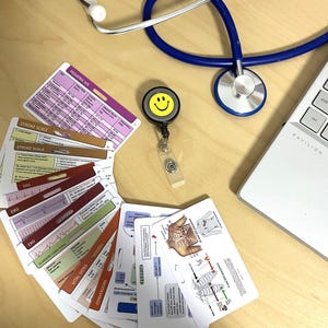 May include: A set of laminated medical reference cards with various medical information, including EKG, stroke scale, Glasgow Coma Scale, vital signs, conversions, and pediatric vital signs. The cards are arranged in a fan shape on a wooden surface. A blue stethoscope and a yellow smiley face badge are also visible in the image.