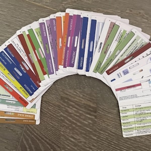 Puede incluir: Un conjunto de tarjetas de referencia médica multicolores dispuestas en abanico. Cada tarjeta presenta una pestaña distintiva codificada por colores con texto impreso que incluye etiquetas como Blood, Therapeutic, Antidotes, Antepartum, Pediatric, Spanish y Pain Scale.
