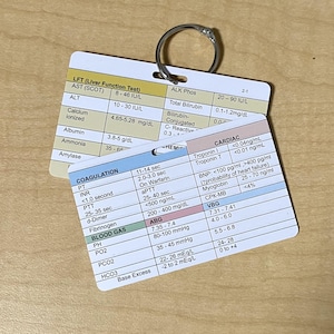 May include: Two laminated medical reference cards with a silver ring. The cards display medical test results, including LFT, ALT, and cardiac markers. The cards are white with black and colored text, providing quick access to vital health information.