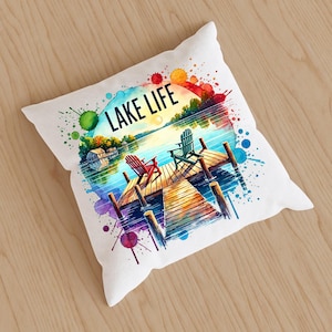 May include: White throw pillow with a watercolor illustration of a lake scene. The illustration features two chairs on a wooden dock, a lake with a sunset, and the text "LAKE LIFE".