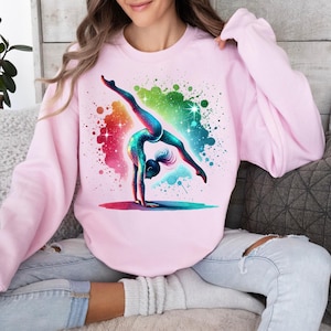 May include: A pink sweatshirt featuring a colourful graphic of a gymnast performing a handstand on a beam. The graphic is surrounded by a colourful splatter paint effect.