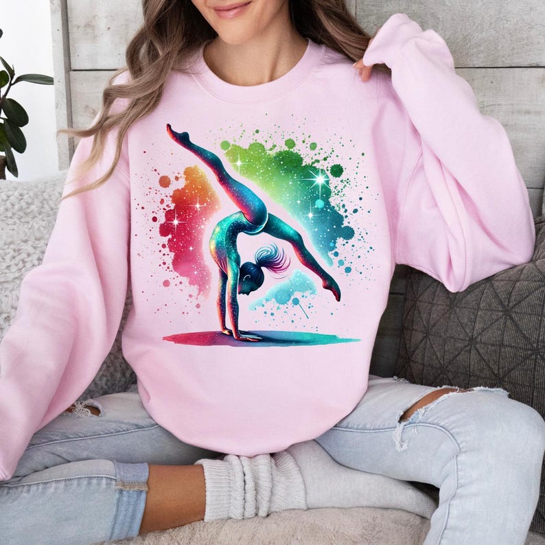 May include: A pink sweatshirt featuring a colourful graphic of a gymnast performing a handstand on a beam. The graphic is surrounded by a colourful splatter paint effect.