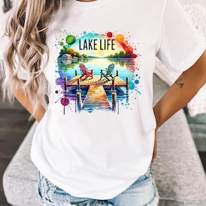 May include: White t-shirt with a watercolor design of a lake scene. The design features two chairs on a dock, a lake with a sunset, and the text "LAKE LIFE".