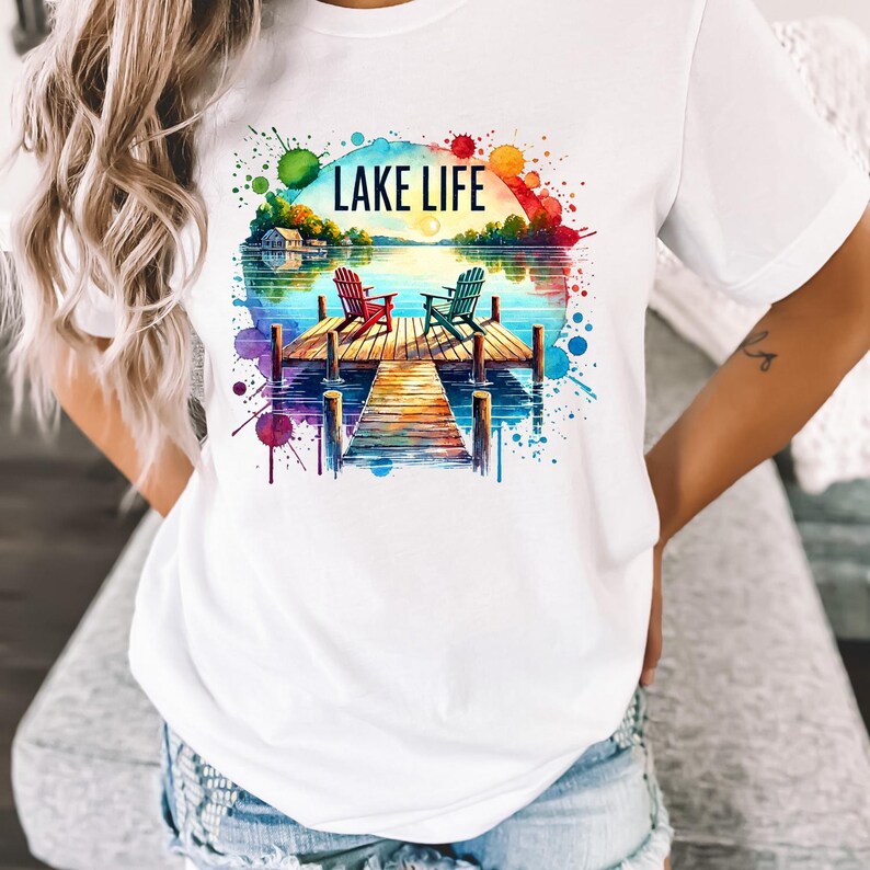 May include: White t-shirt with a watercolor design of a lake scene. The design features two chairs on a dock, a lake with a sunset, and the text "LAKE LIFE".