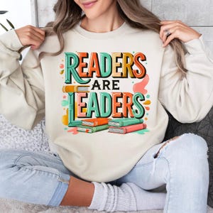 May include: Cream-colored sweatshirt with the text "READERS ARE LEADERS" in a colorful, 3D design. The graphic includes stacks of books in orange, green, and red. A casual, comfortable top for book lovers and educators.